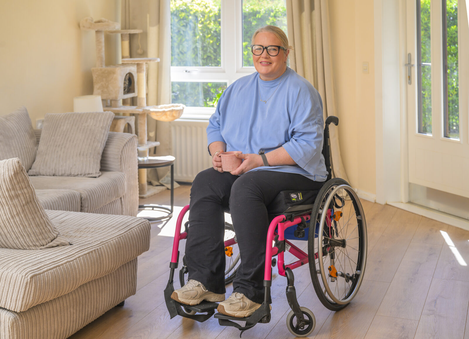 A woman in a wheelchair in her living room, holding a mug of tea and smiling